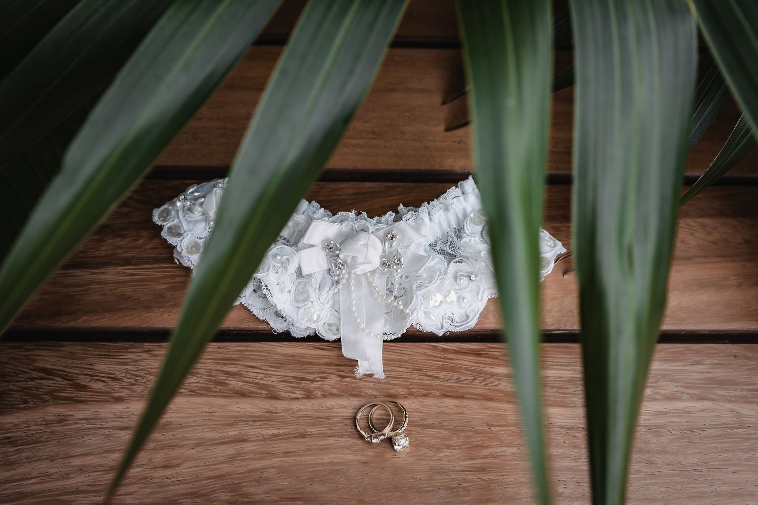 White lace wedding garter and gold rings displayed on rustic wooden floor with tropical green leaves.