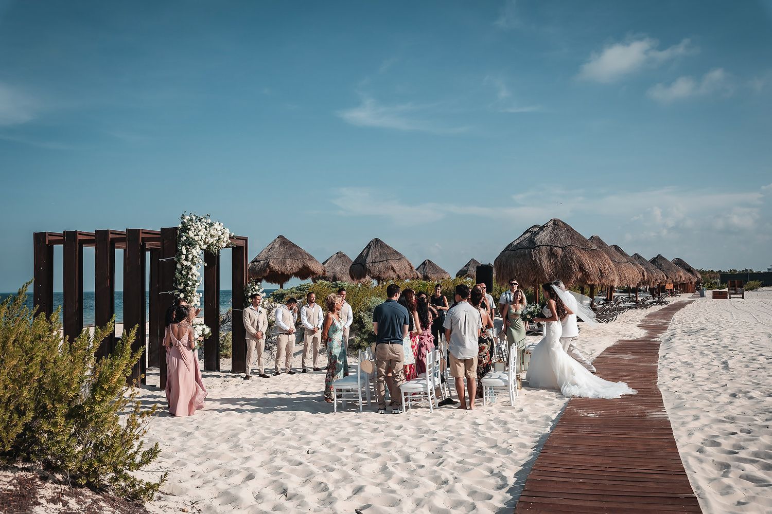 Beach wedding ceremony with guests gathered around a floral arch on white sand, tropical thatched huts in the background.