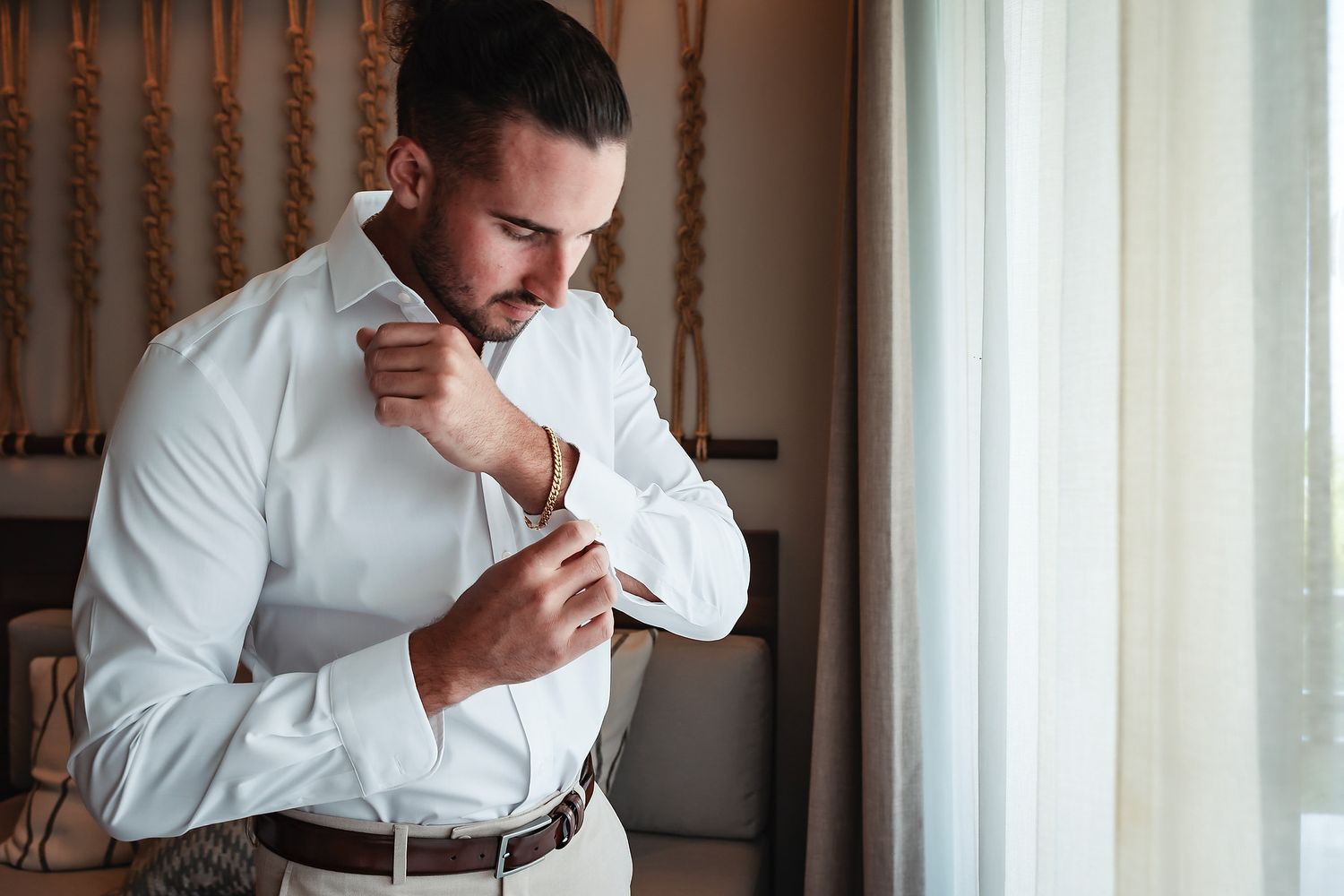 A well-dressed man in a white dress shirt fastening his cufflinks while standing near a bright window.