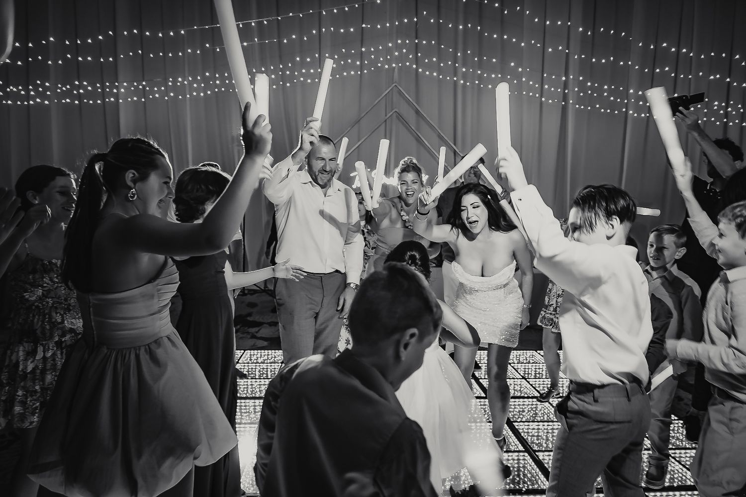 Black and white photo of wedding guests dancing joyfully with glow sticks raised at a festive indoor reception.
