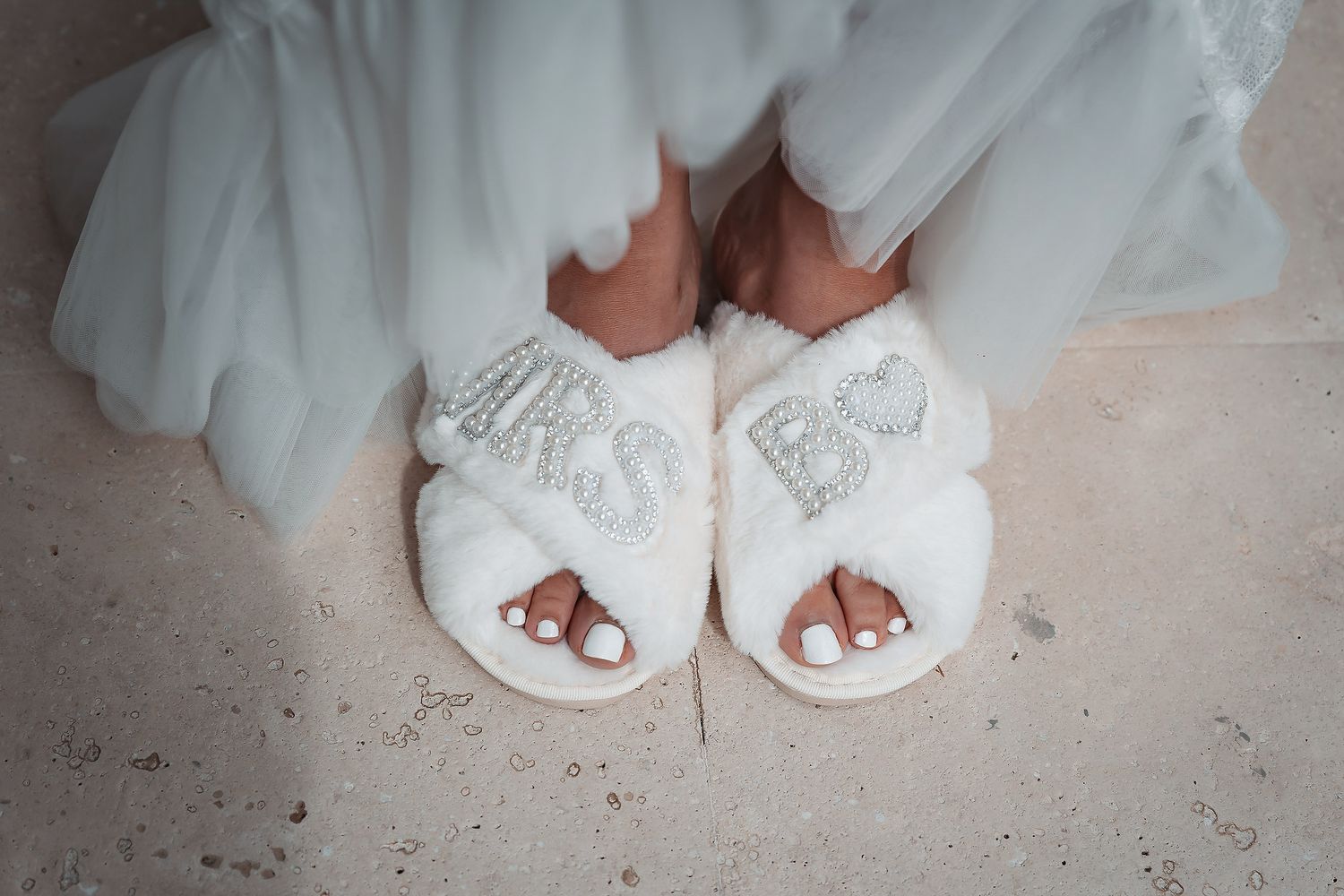Bride wearing white fluffy MRS slippers with rhinestone letters on marble floor.