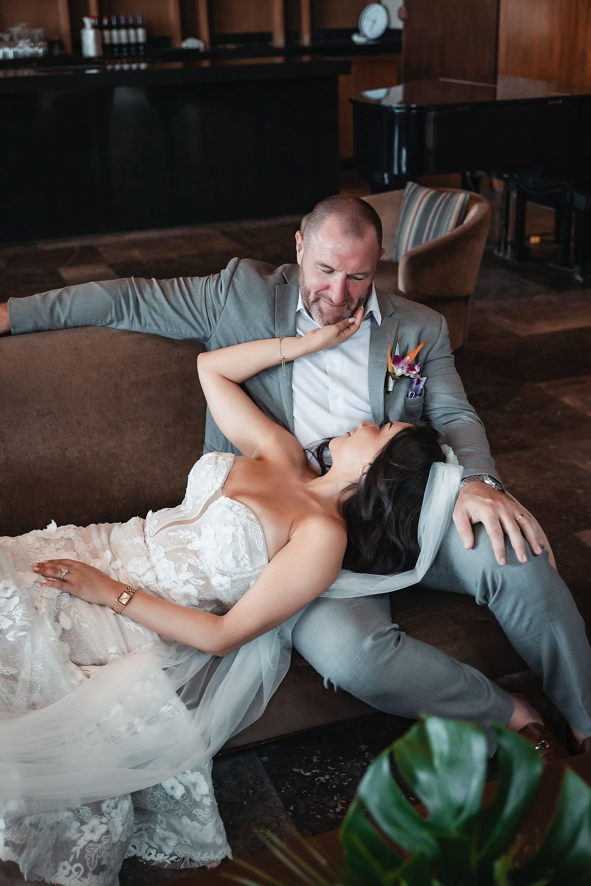 Bride reclines across grooms lap on a couch, sharing a joyful moment in a warmly lit wedding venue.