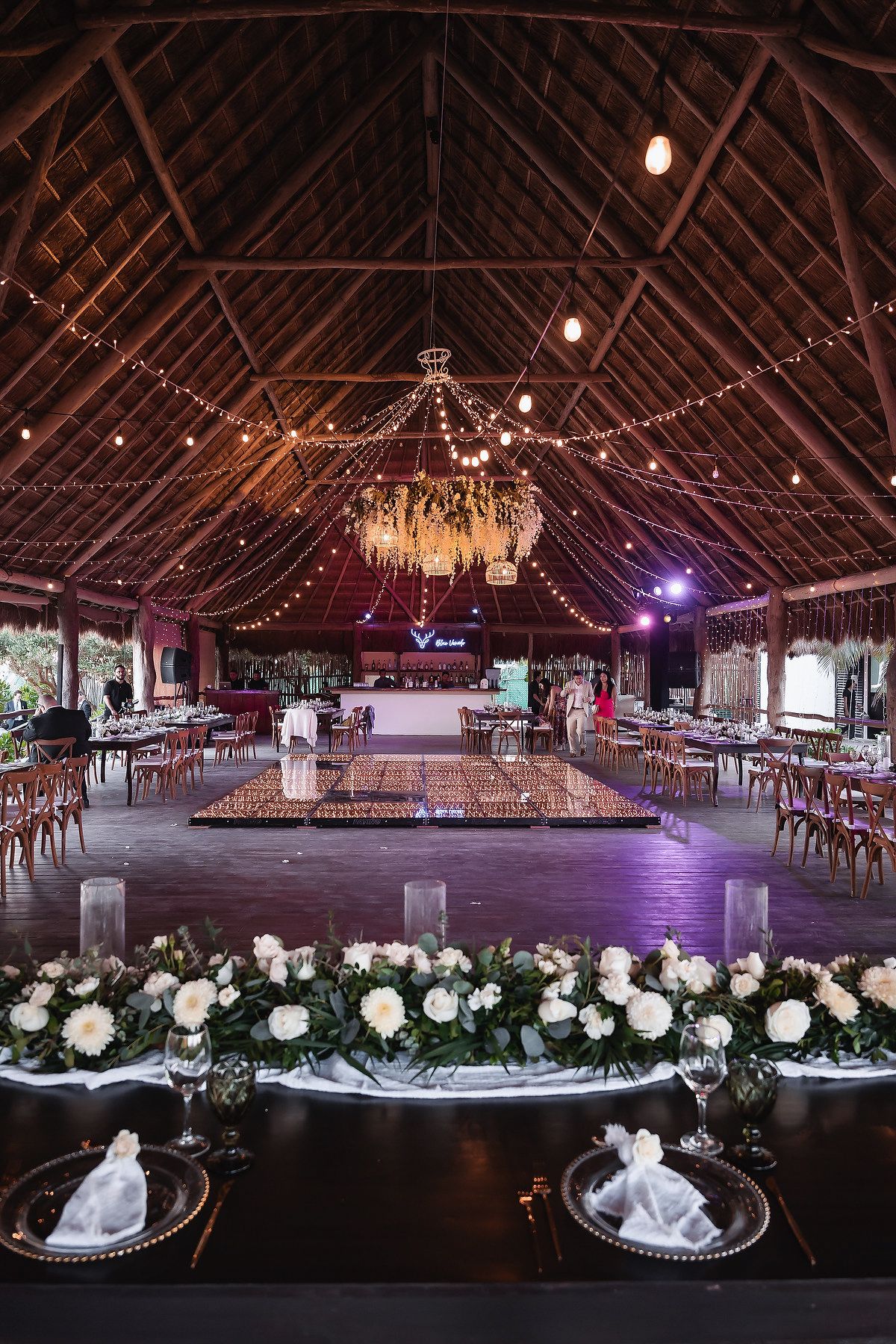 Elegant wedding reception venue with string lights, chandelier, dance floor, white floral centerpieces, and rustic wood-beamed ceiling.