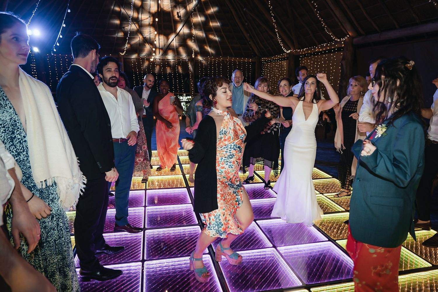 Guests dancing on illuminated LED dance floor at wedding reception under thatched roof venue.