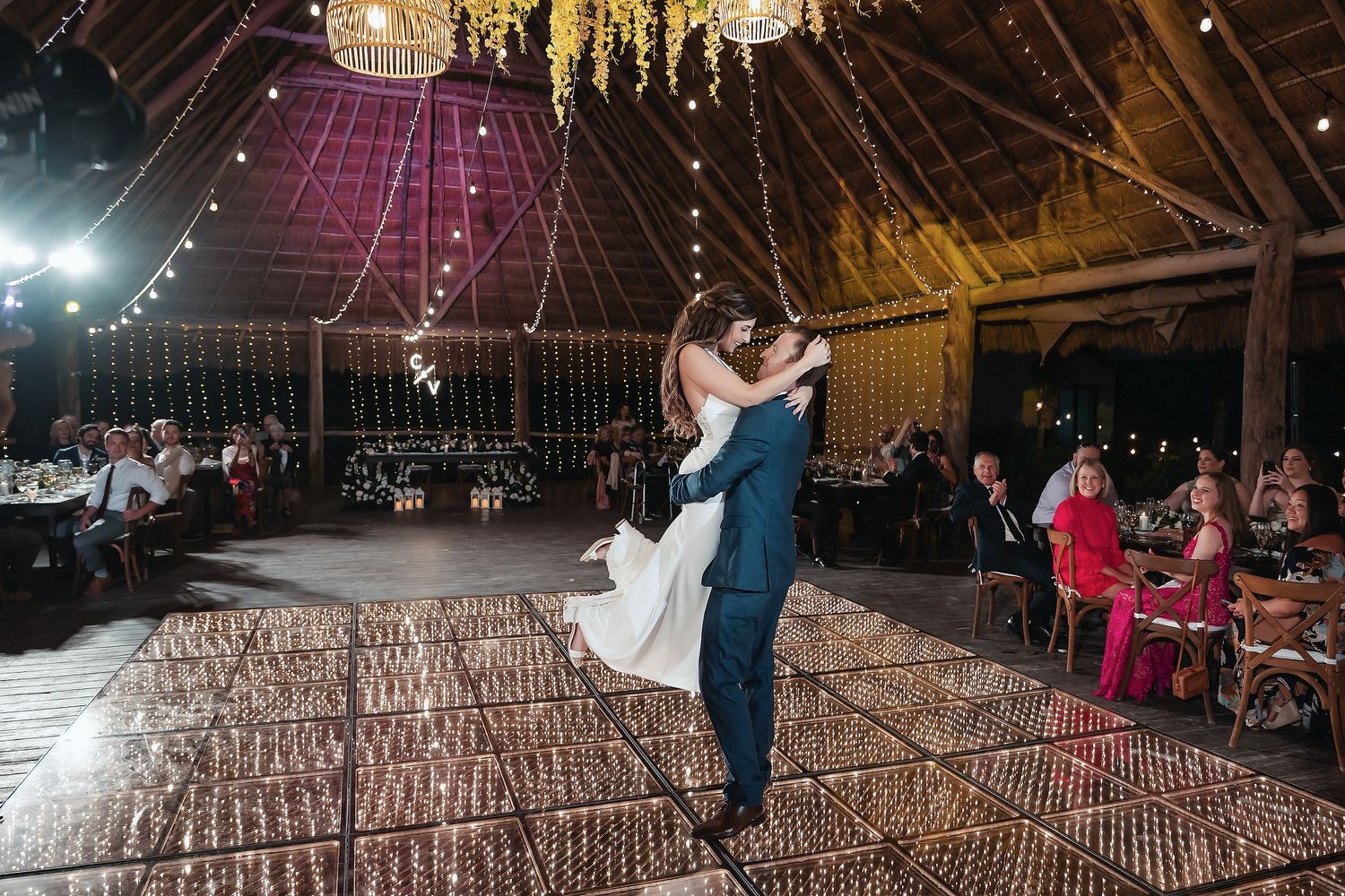 Groom lifts bride during first dance in a rustic barn wedding venue with string lights and floral decor.