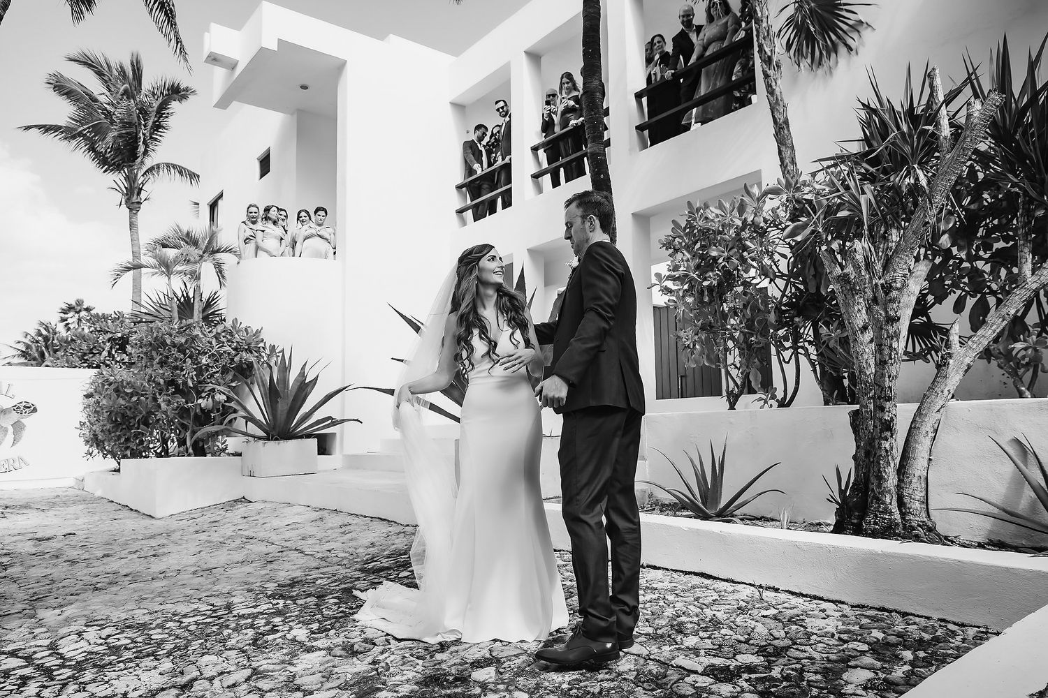 Black and white photo of a couple sharing a romantic moment at an outdoor wedding venue with tropical plants.