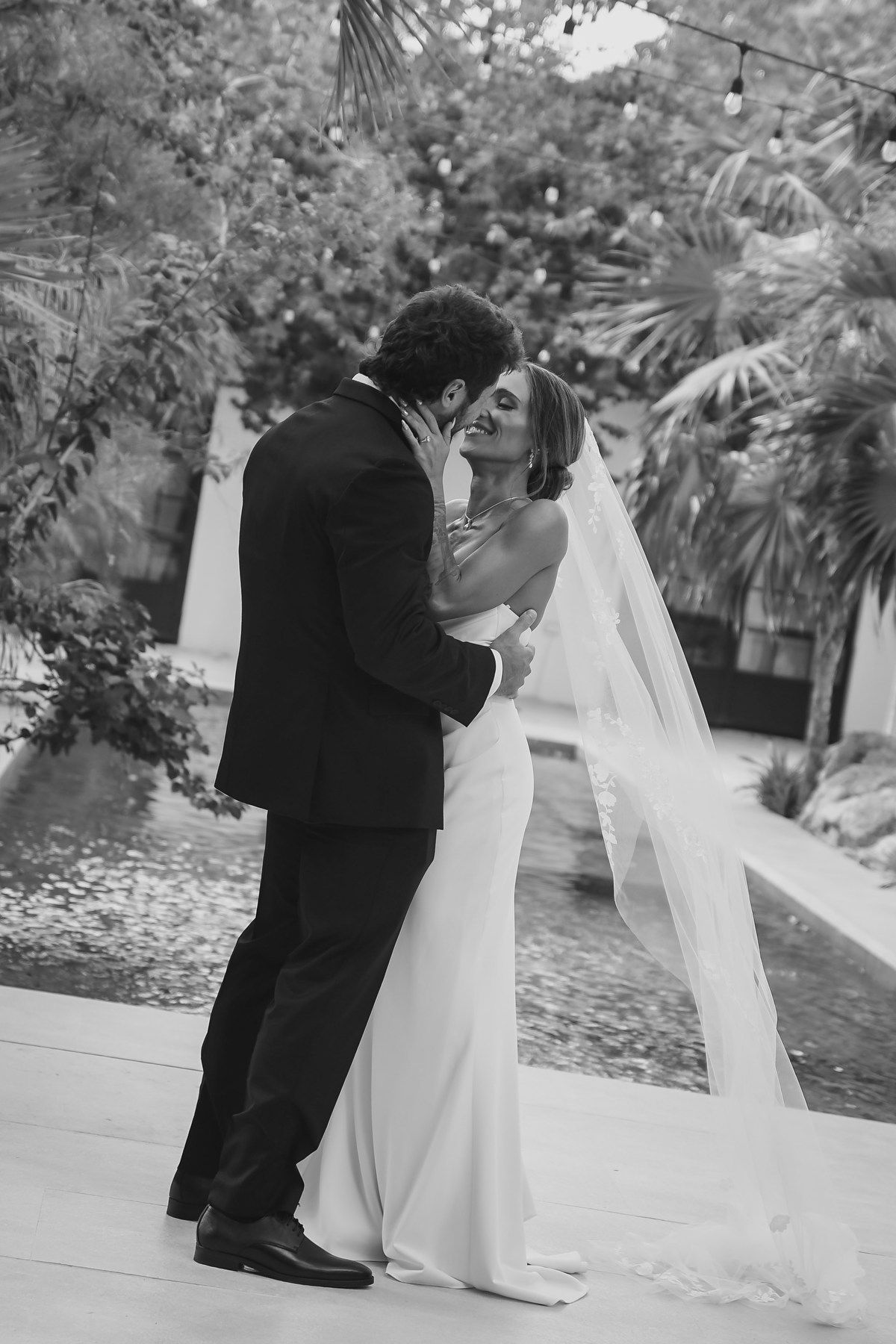 Black and white photo of bride and groom sharing a romantic moment by a pool surrounded by tropical trees.