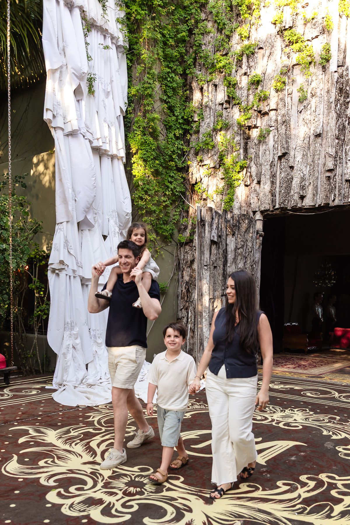 A happy family of four walks through a lush, artistic outdoor space with rock walls and decorative flooring.