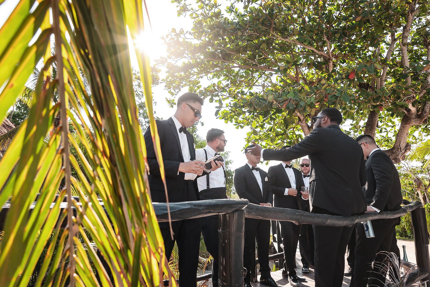 Groomsmen in black tuxedos gather outdoors under lush trees in golden sunlight before a wedding ceremony.