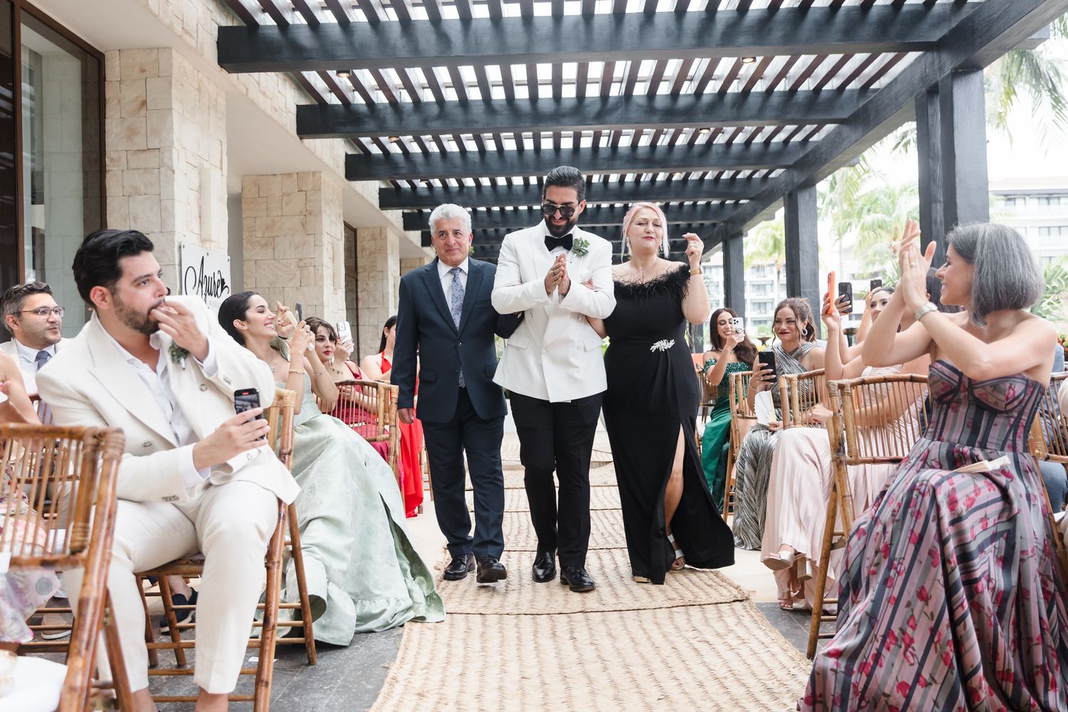 Guests gather for an outdoor wedding ceremony under a pergola, with the wedding party walking down the aisle.