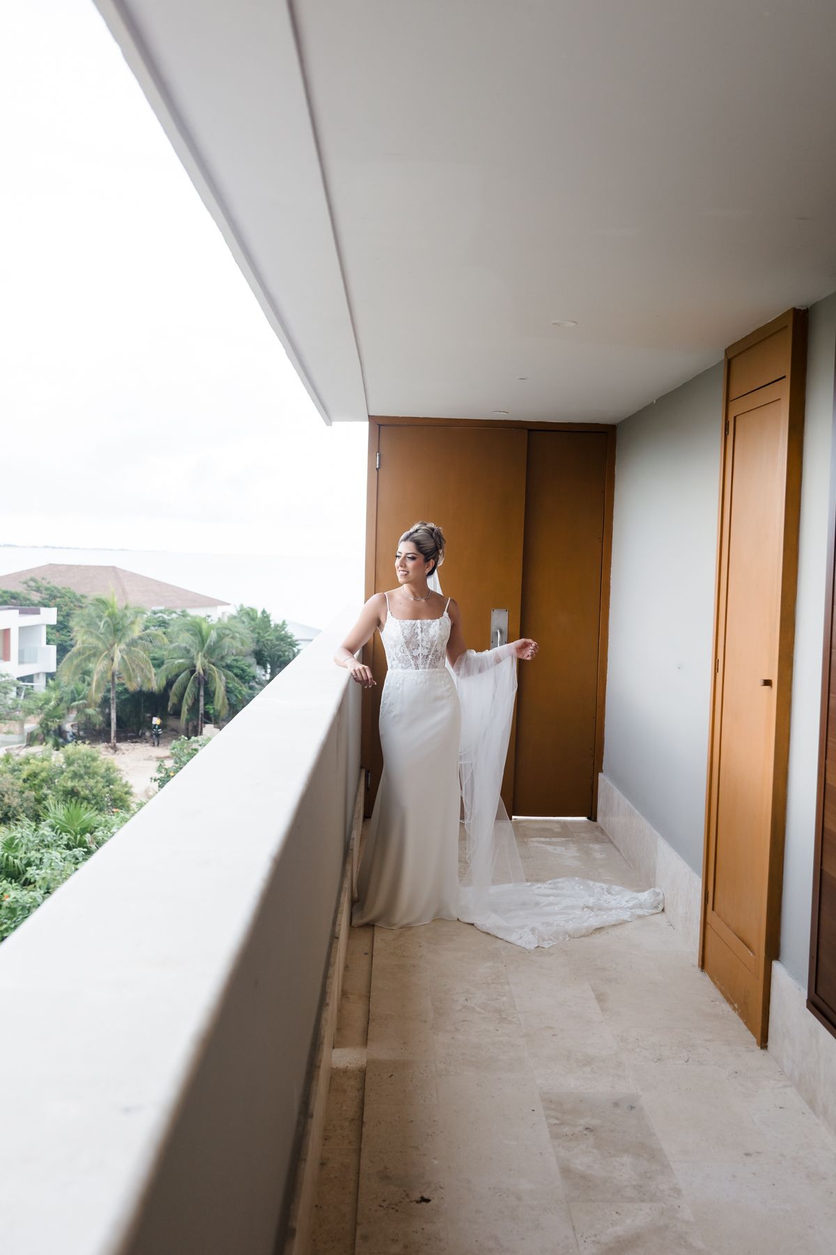 Bride in white gown with veil stands on hotel balcony overlooking tropical palms on her wedding day.