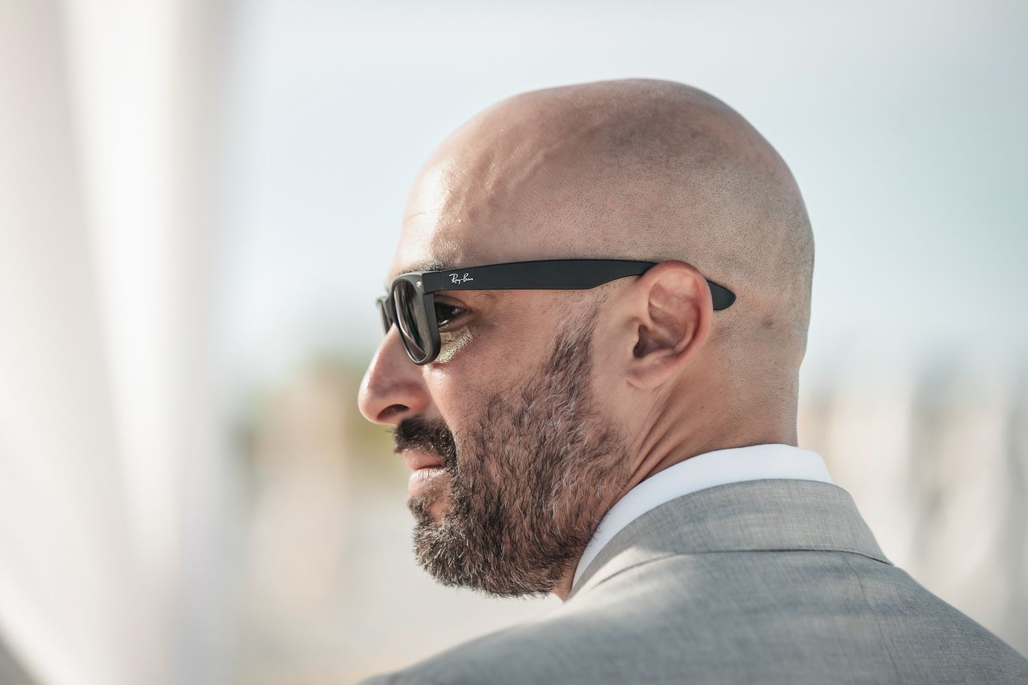 Bald man with beard wearing Ray-Ban sunglasses and gray suit jacket, photographed in profile outdoors.