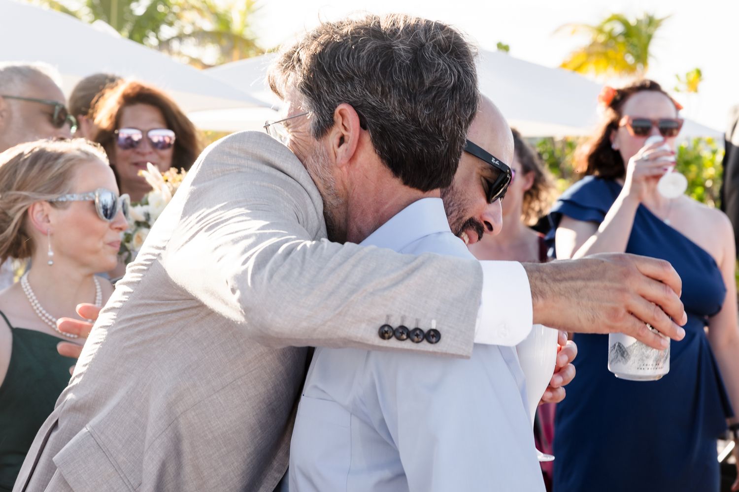 Two men share a warm hug at an outdoor wedding reception, with guests in formal attire visible in the background.