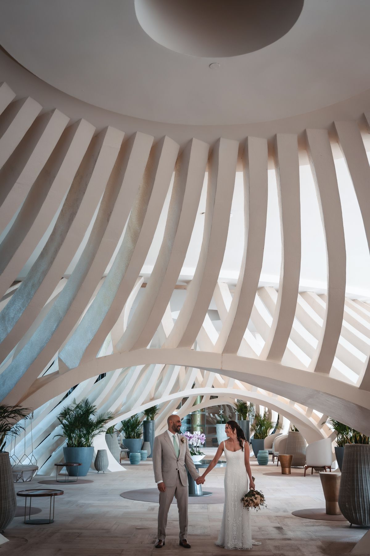 Bride and groom holding hands under stunning curved white architectural ribs in an elegant resort lobby.
