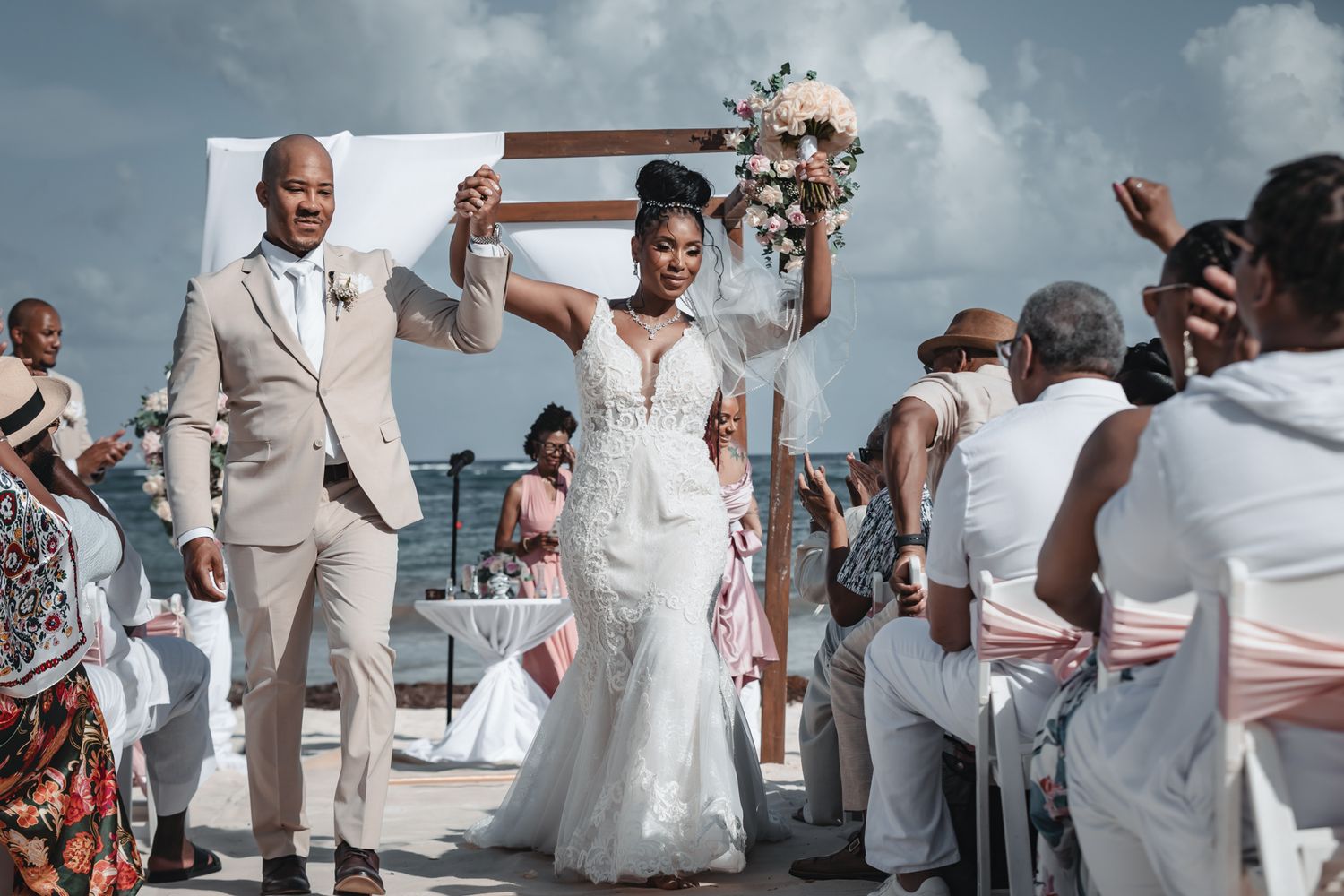 Bride and groom celebrate at outdoor beach wedding ceremony, bride raises bouquet joyfully.