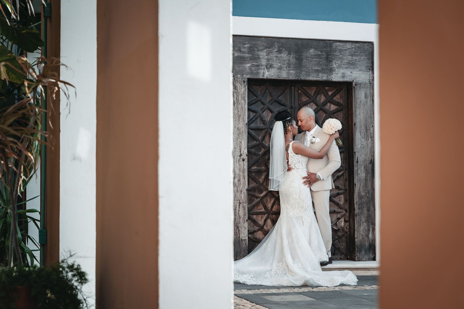 Bride and groom share a romantic kiss in white wedding attire before ornate wooden doors at an elegant outdoor venue.