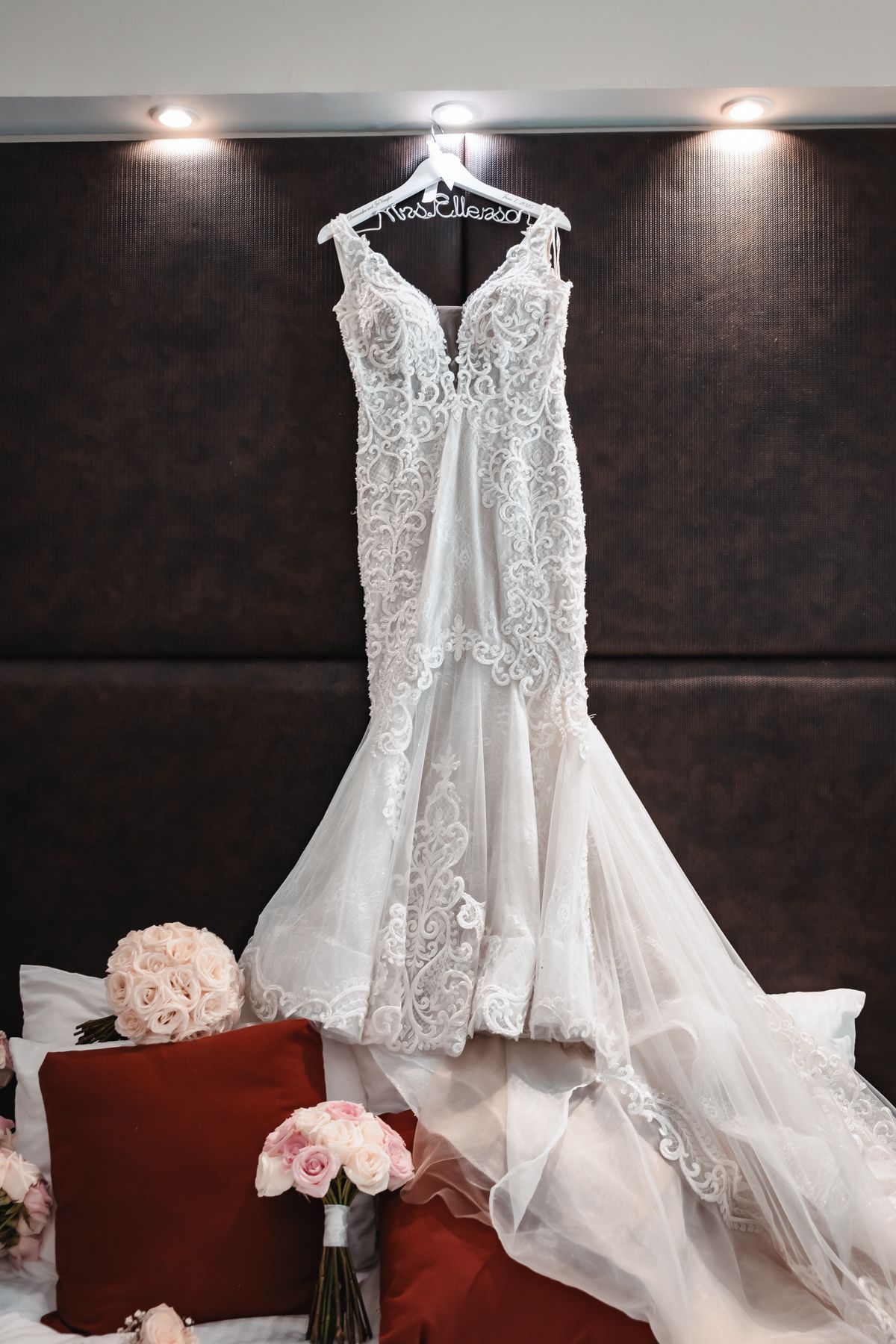 Elegant white lace mermaid wedding dress hanging on a bridal hanger, surrounded by pink roses on a bed.