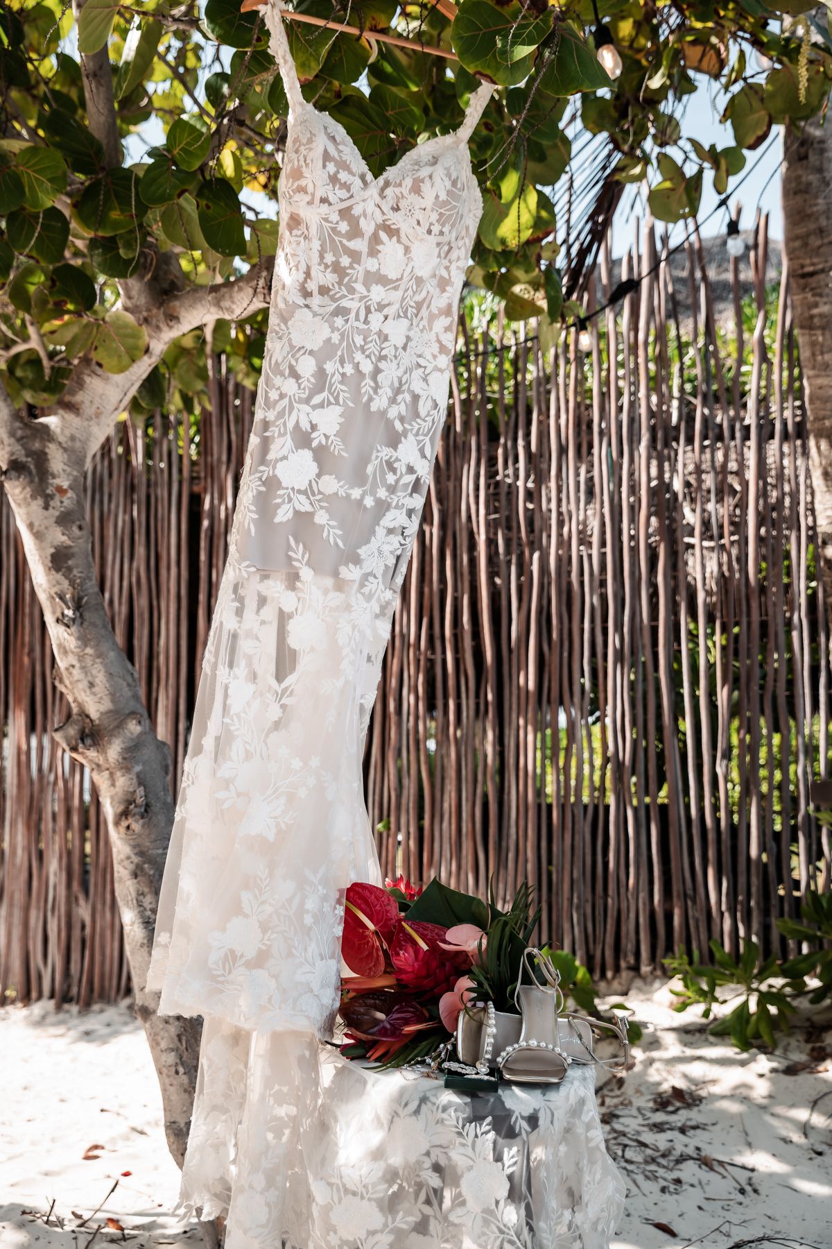 Elegant lace wedding dress hanging from a tree on a sandy beach with a red floral bouquet.