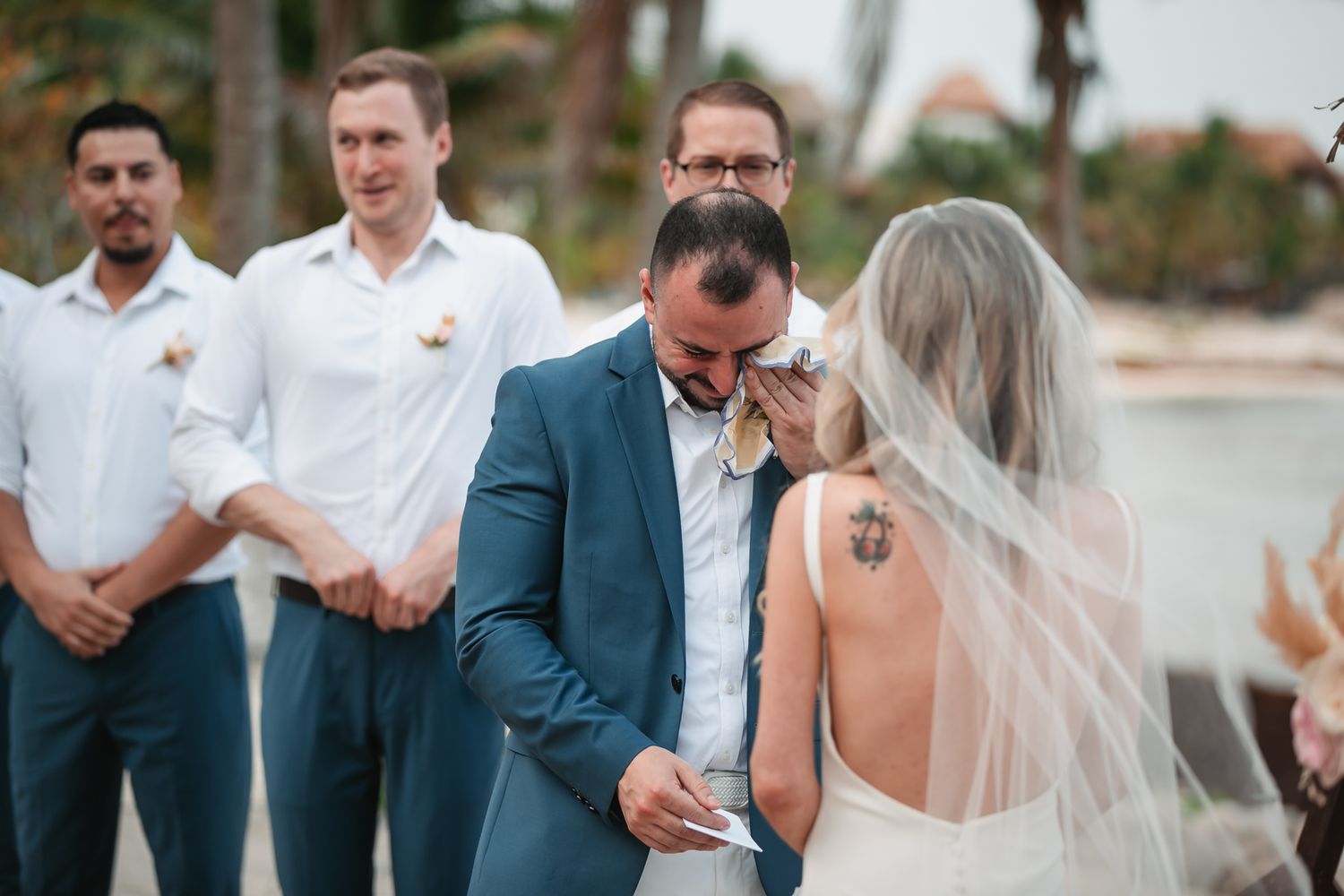 Emotional groom wiping tears reading vows to bride during outdoor beach wedding ceremony.