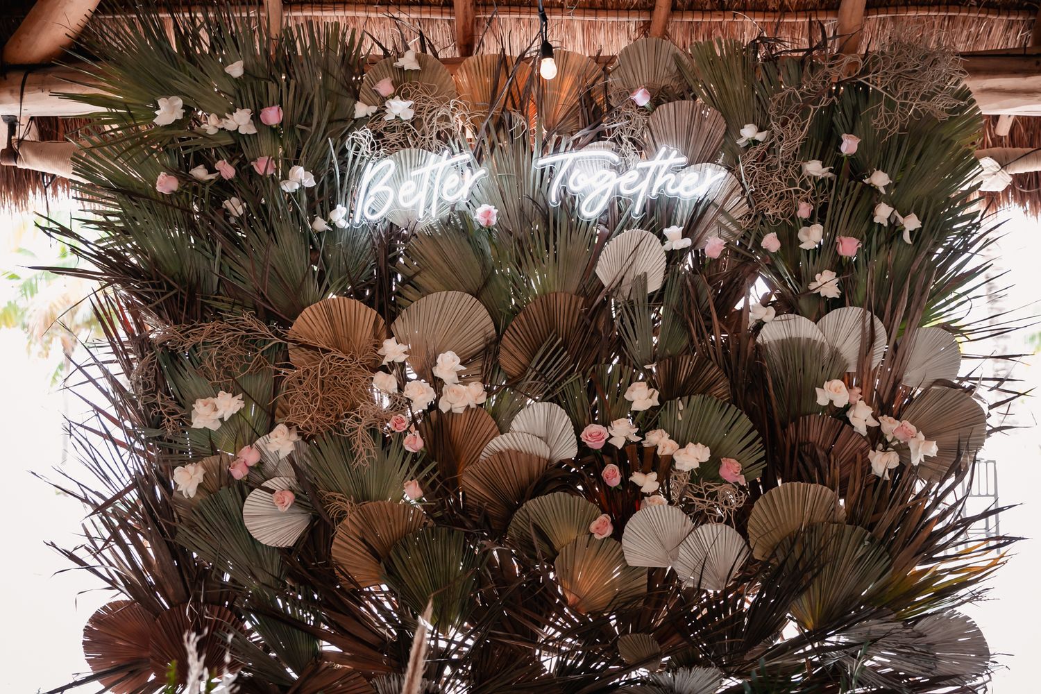 Boho wedding backdrop with dried tropical leaves and pink flowers featuring Better Together neon sign.