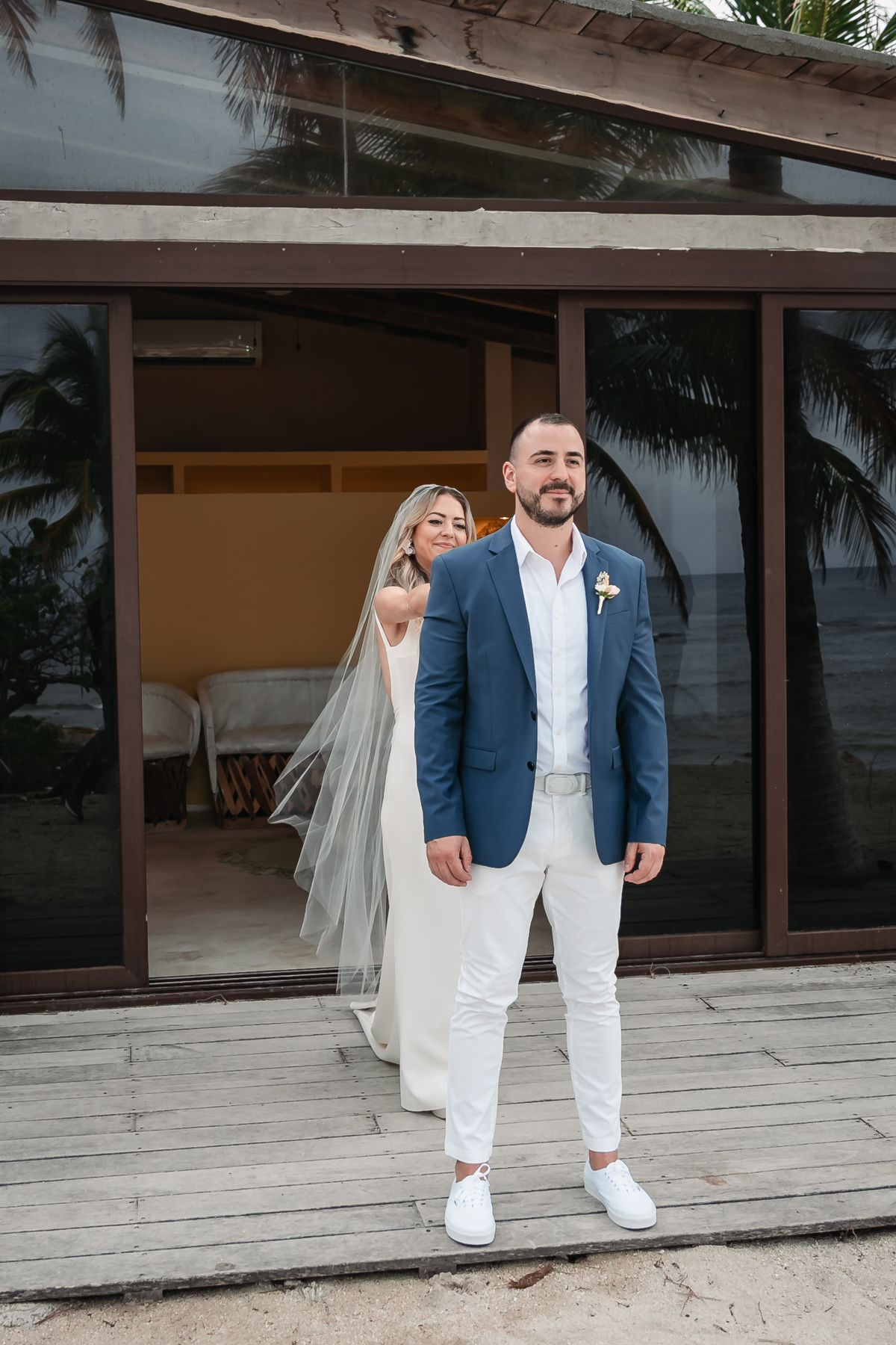 Bride and groom standing on a wooden deck outside a tropical beach villa surrounded by palm trees.