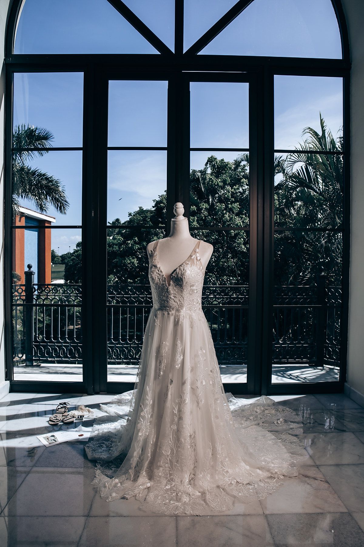 Three elegant white wedding dresses displayed in a sunlit room with large windows and palm trees visible outside.