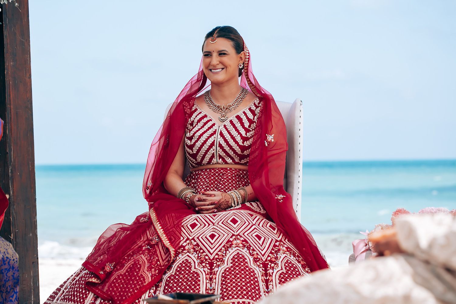Bride in a stunning red and white embroidered lehenga poses gracefully on a beach with turquoise waters in the background.