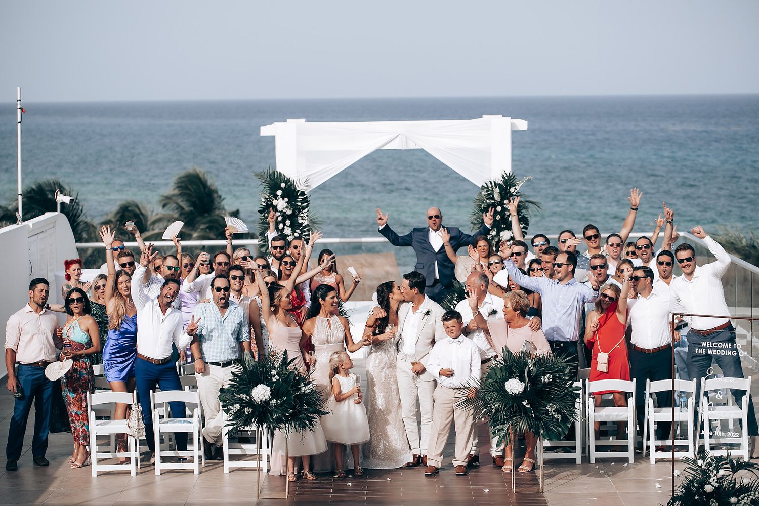 Wide shot of a seaside wedding ceremony with guests seated in white chairs and tropical greenery arrangements.