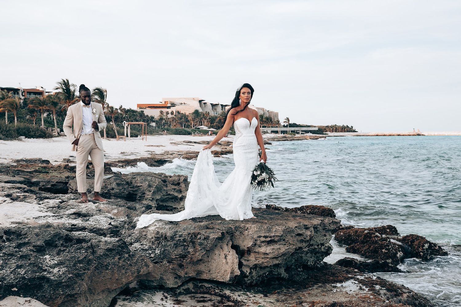 Newlyweds pose on rocky oceanfront with coastal buildings in background.