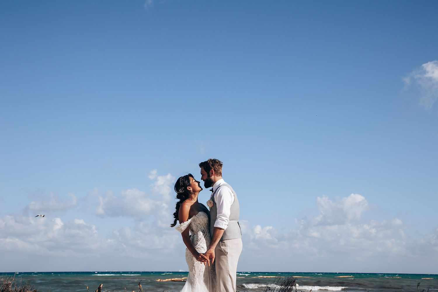 Newlyweds share intimate moments on a sunny beach with waves in the background.