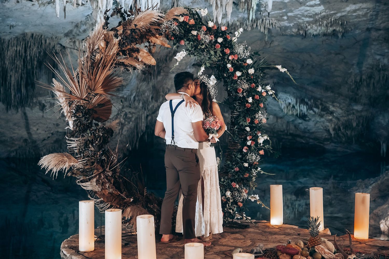 A romantic wedding ceremony setup in a cave with a floral arch, pampas grass, and glowing candles on the ground.