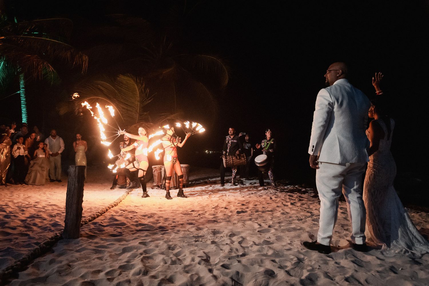 Fire dancers performing spectacular nighttime beach show for Indian wedding couple and guests on sand.