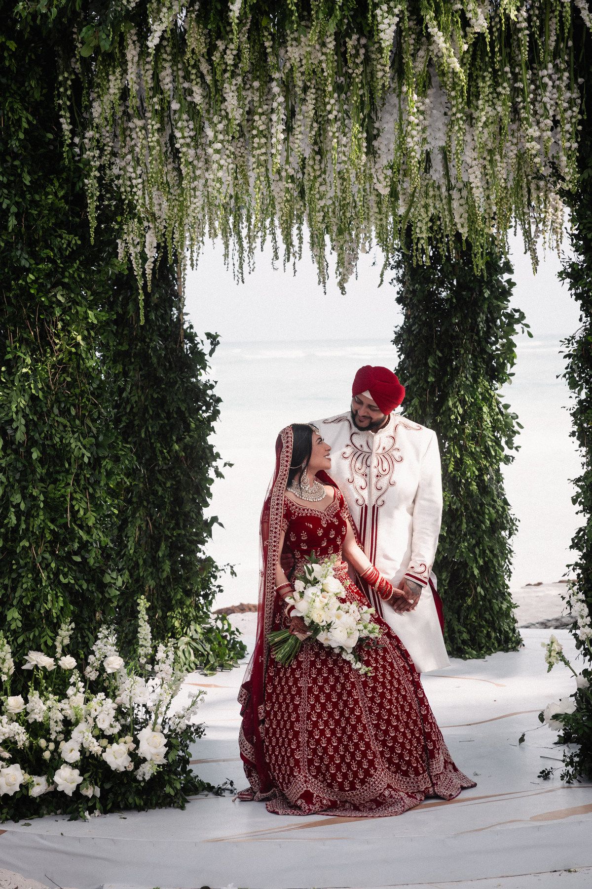 Indian bride and groom pose under floral arch with white flowers at elegant outdoor beach wedding.