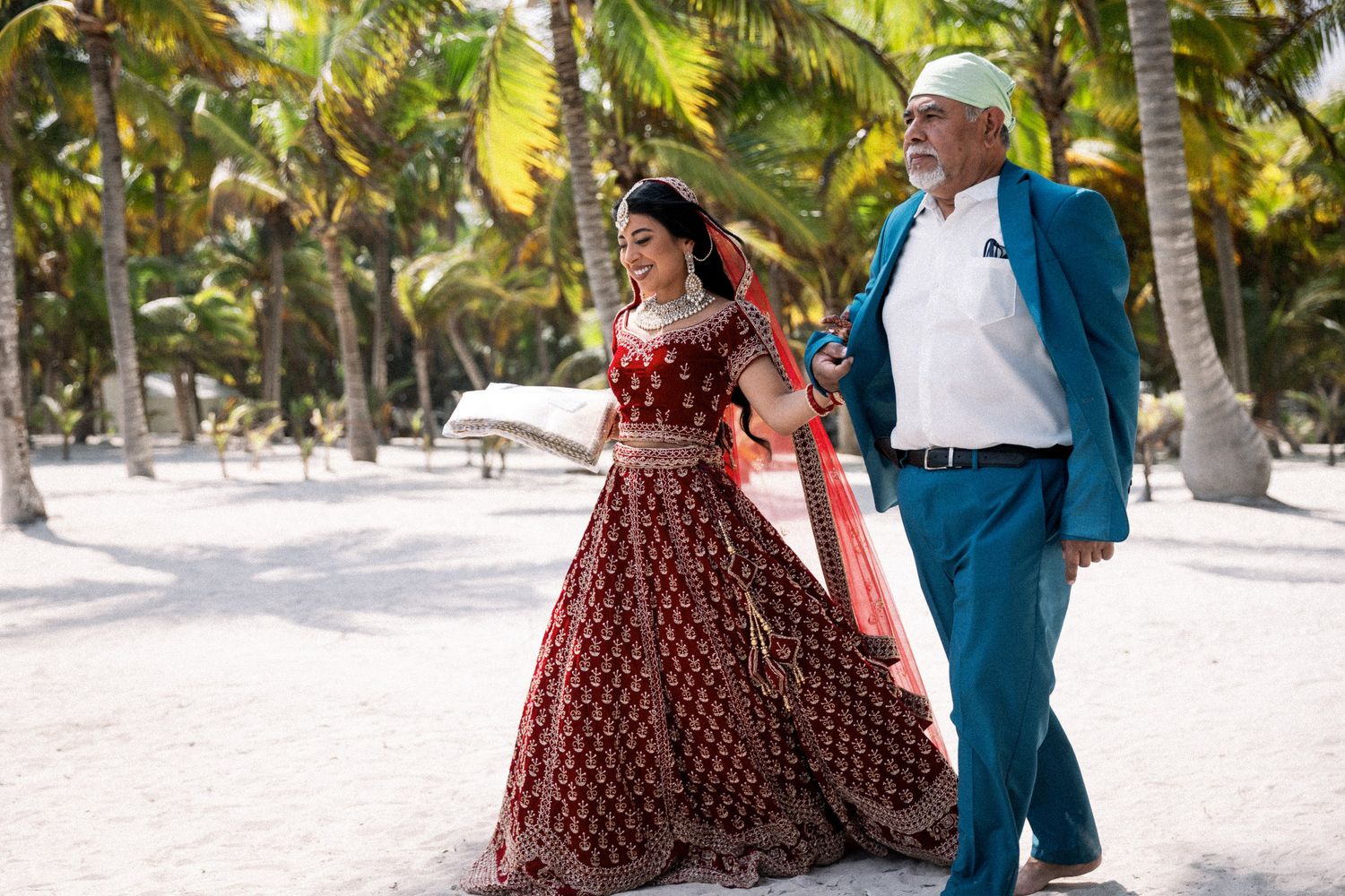 Indian bride in red lehenga walking with father on sandy beach surrounded by tropical palm trees.