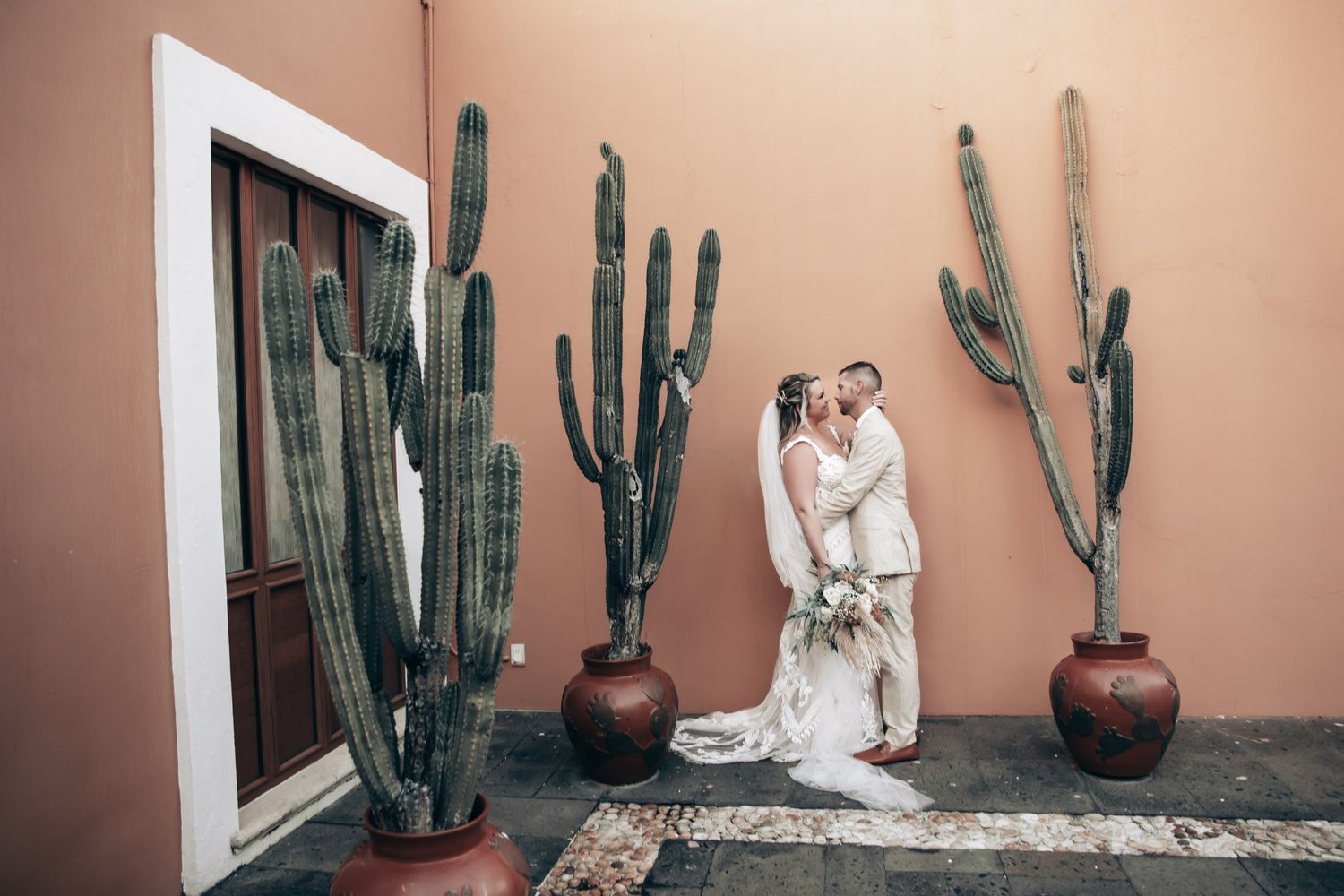 Bride and groom share a romantic kiss surrounded by tall cacti against a warm terracotta wall at Dreams Tulum