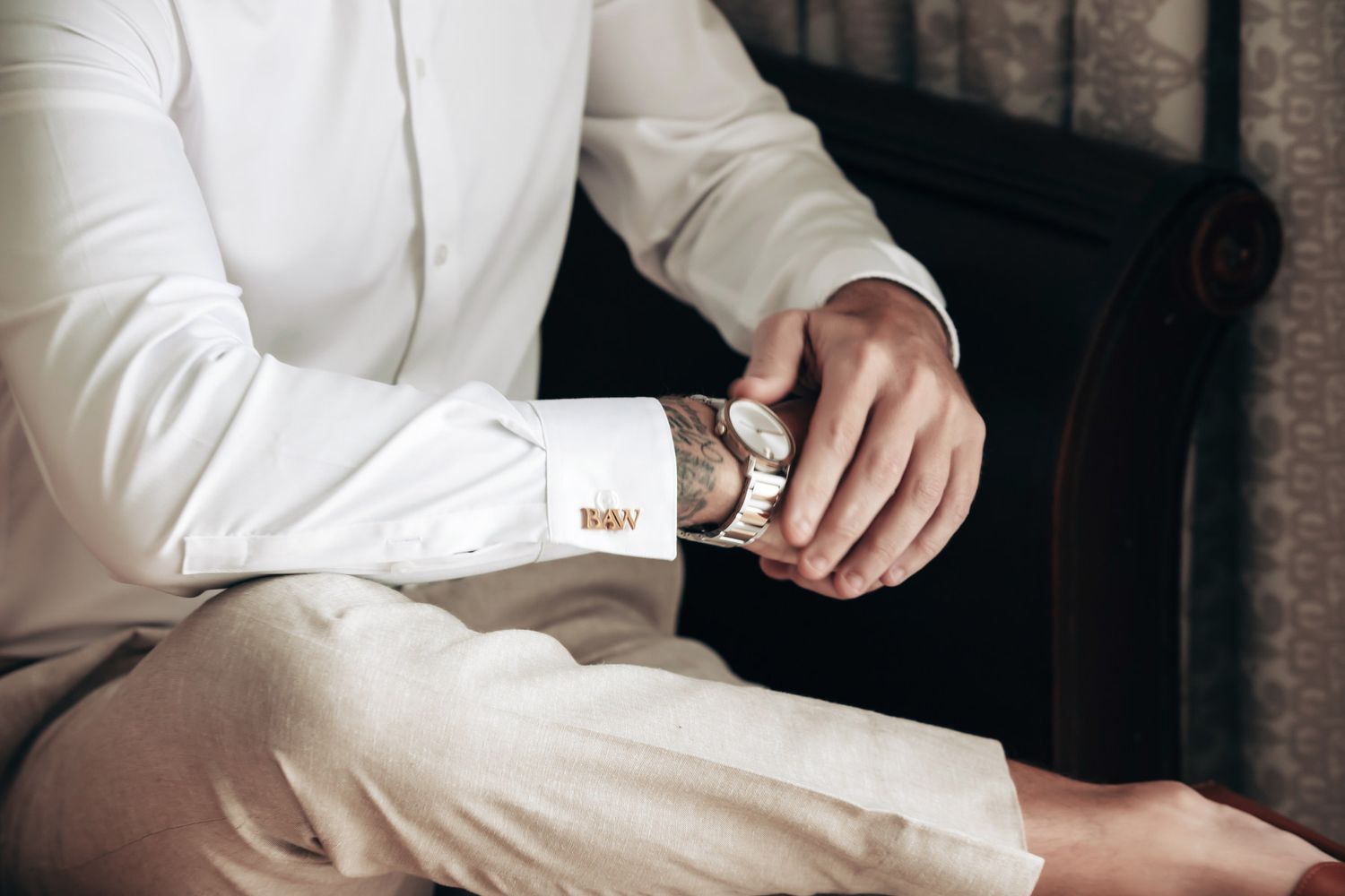 Groom fastening gold cufflinks on white dress shirt sleeve while seated before wedding ceremony.