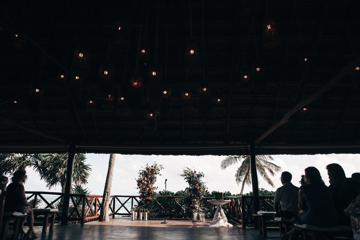Romantic wedding ceremony venue with string lights and silhouetted guests seated by a tropical oceanfront.
