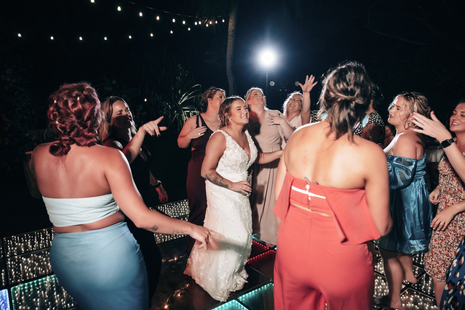 Bride dancing with guests at outdoor wedding reception at night, surrounded by women in colorful dresses under string lights.