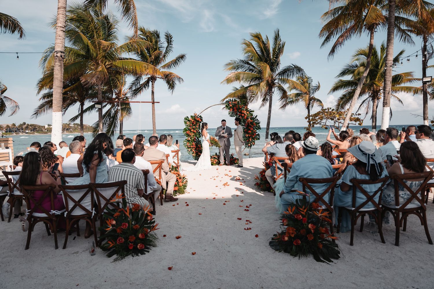 Outdoor beach wedding ceremony with guests seated among tropical palm trees.