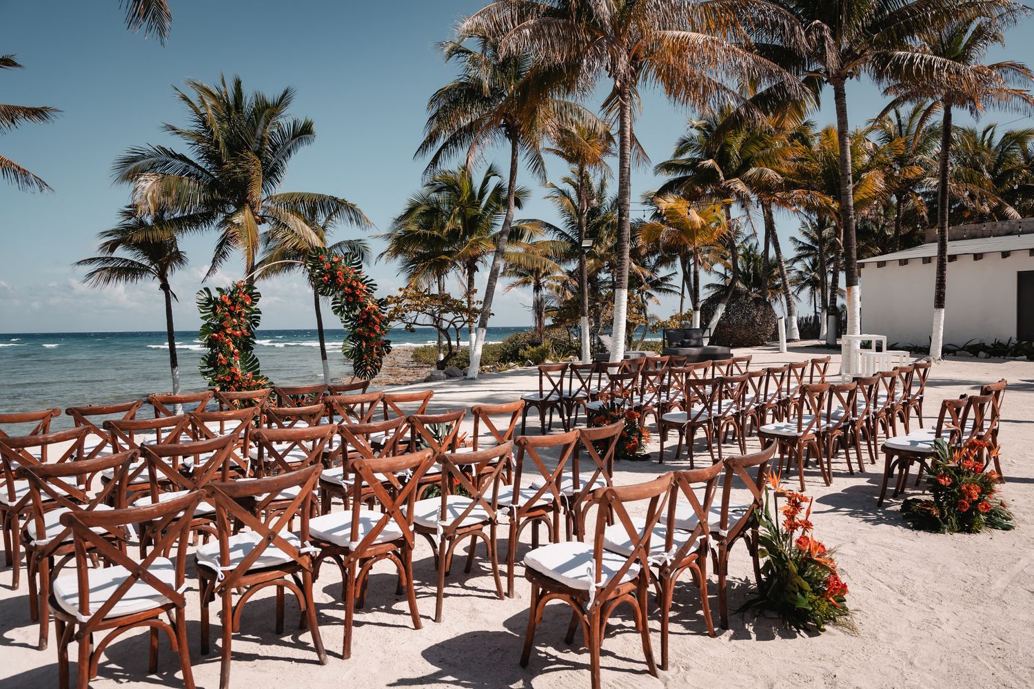 Outdoor beach wedding ceremony setup with wooden cross-back chairs, palm trees, and ocean views under a clear blue sky.