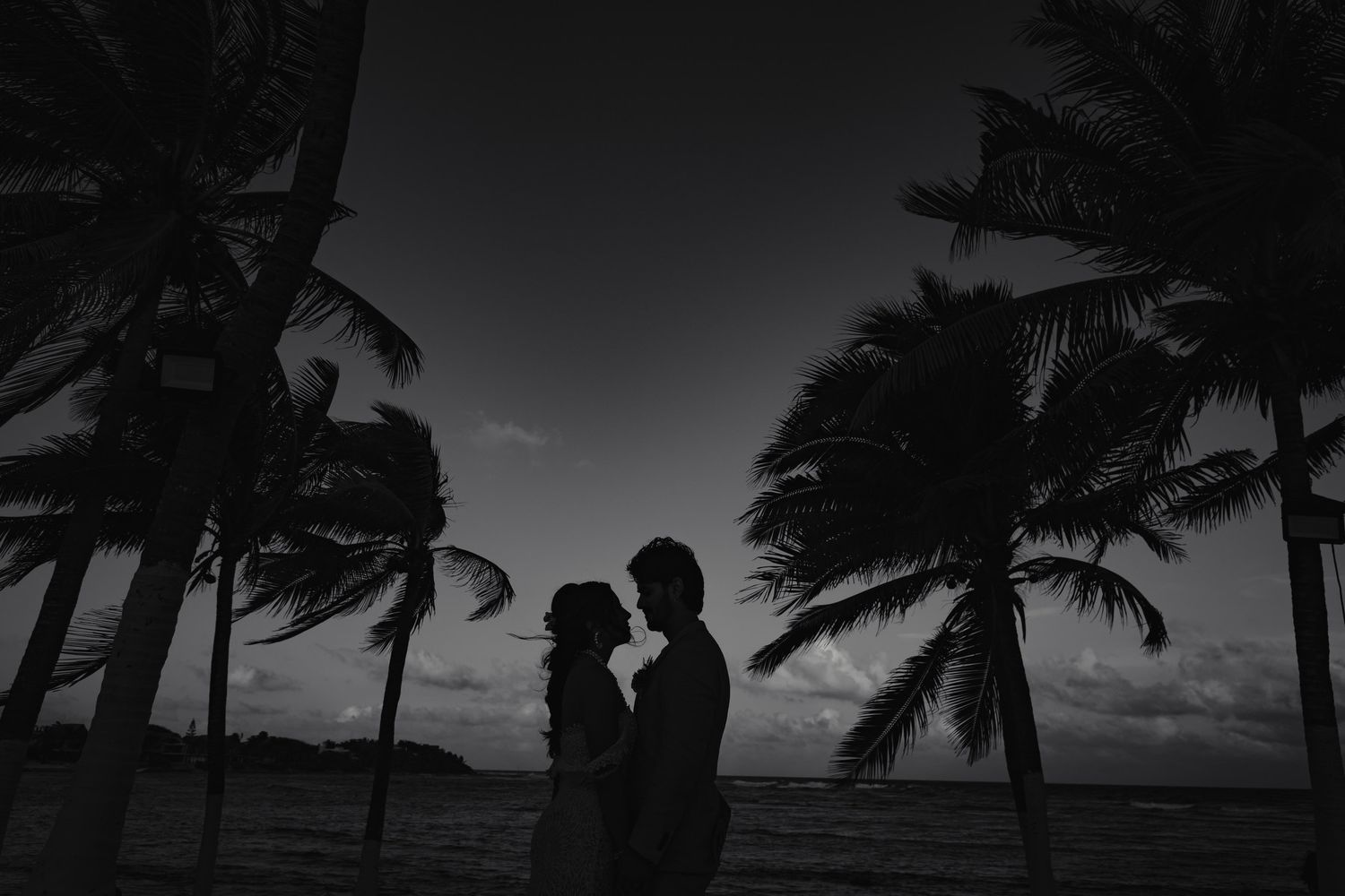 Silhouette of bride and groom at sunset among swaying tropical palm trees.