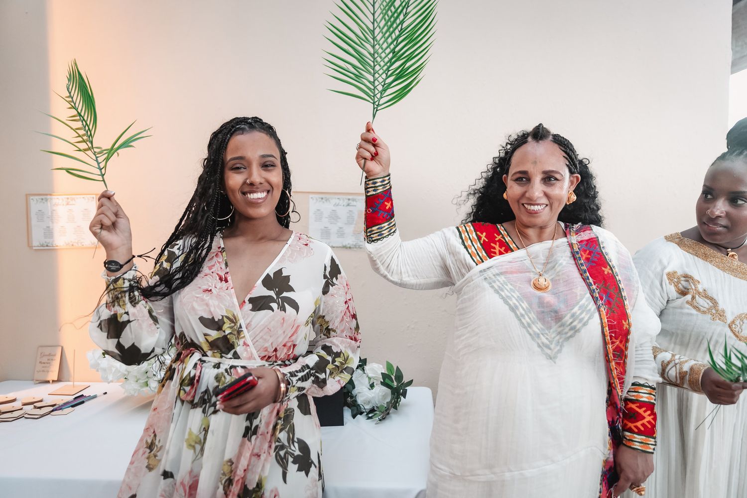 Two women in colorful attire smiling and holding palm fronds during a cultural wedding celebration.