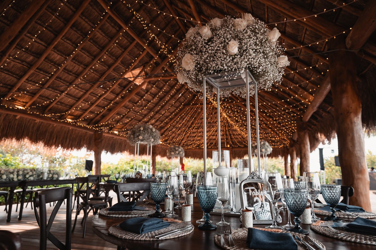 Elegant wedding reception under a thatched palapa with fairy lights and floral chandeliers.
