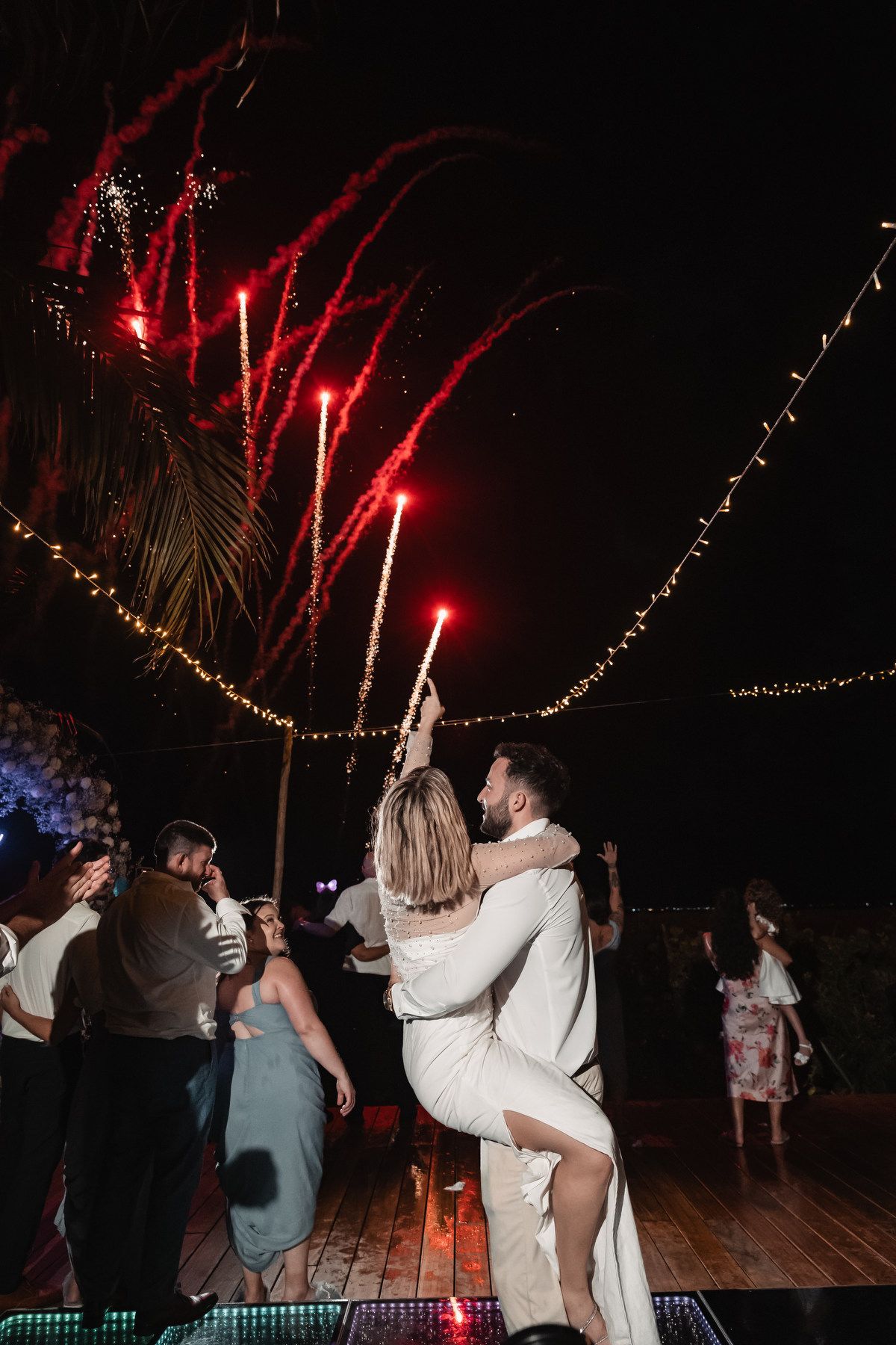 Groom lifting bride as red fireworks light the night sky during outdoor wedding dance celebration.