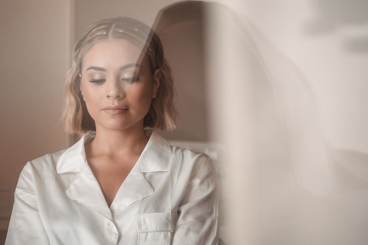 Young woman with blonde hair in white satin pajamas posing with eyes closed in soft natural light.