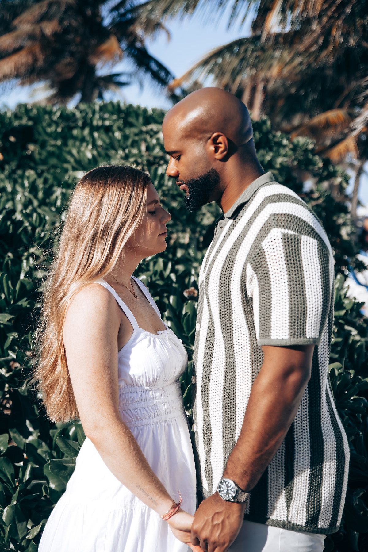 Interracial couple holding hands and gazing at each other near lush tropical greenery and palm trees.