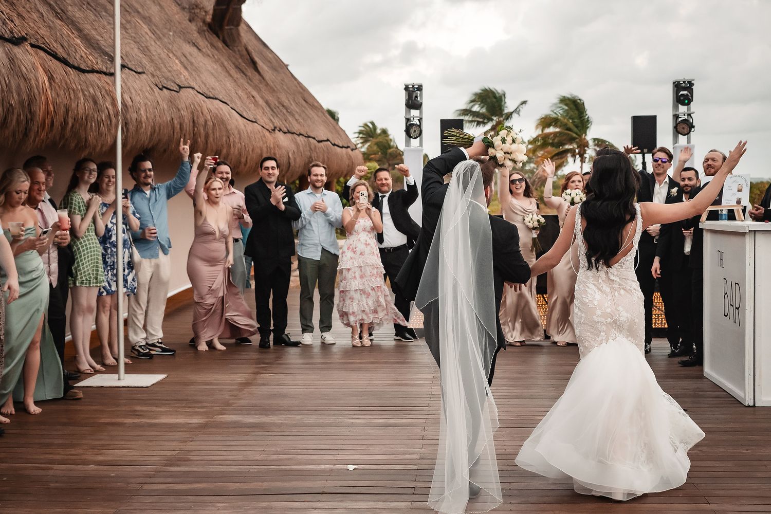 Bride and groom share first dance on tropical resort deck while wedding guests celebrate around them.