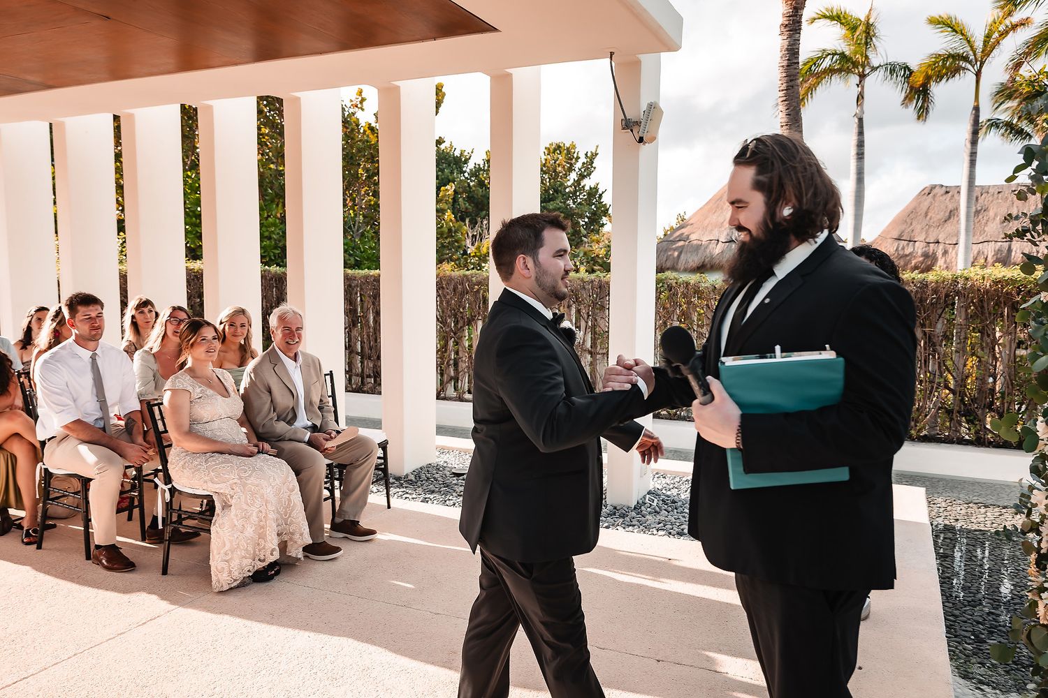 Two grooms exchange vows at an outdoor wedding ceremony with guests seated nearby, surrounded by tropical palm trees.