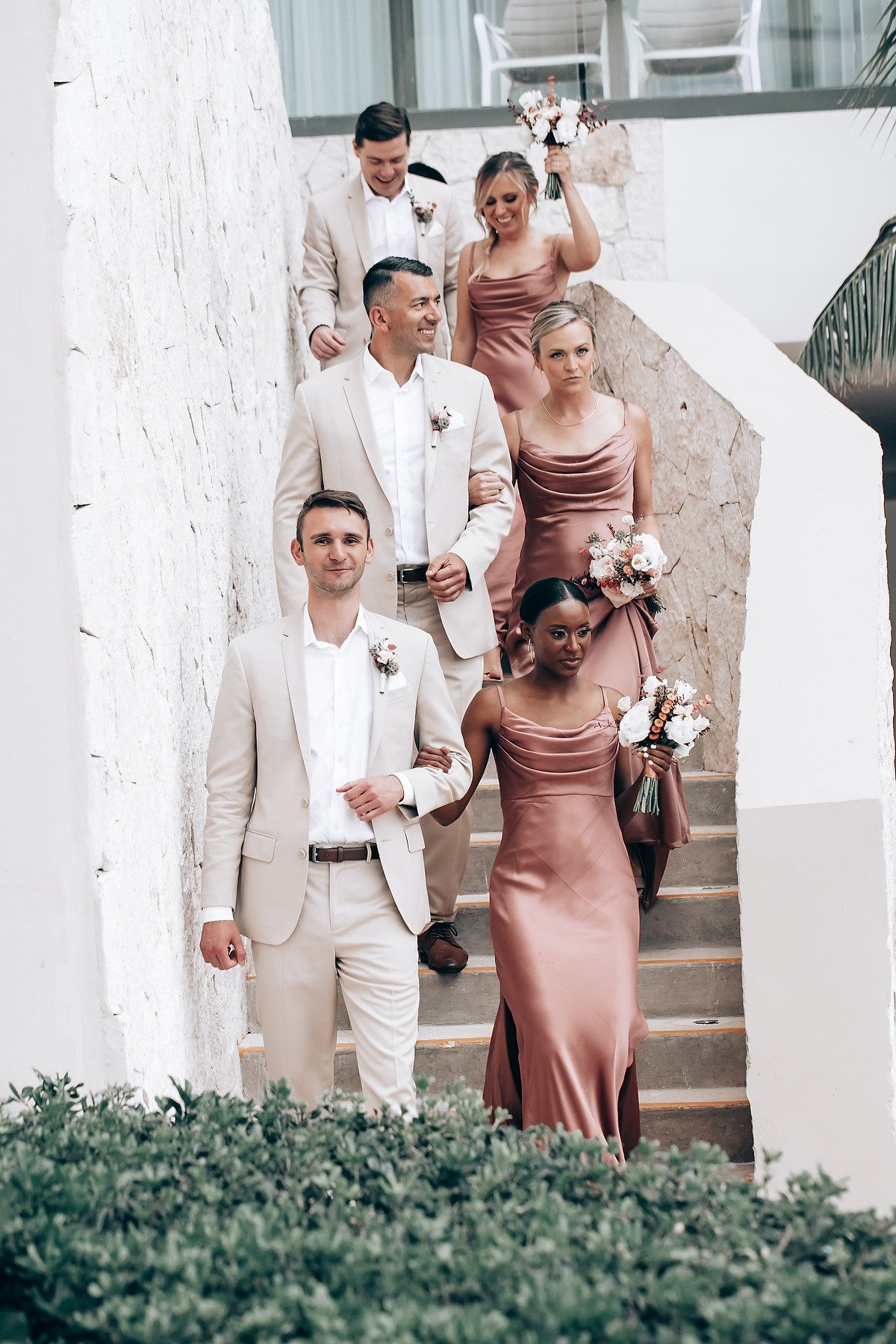Wedding party members in light beige suits and dusty rose dresses stand together on white columned steps.