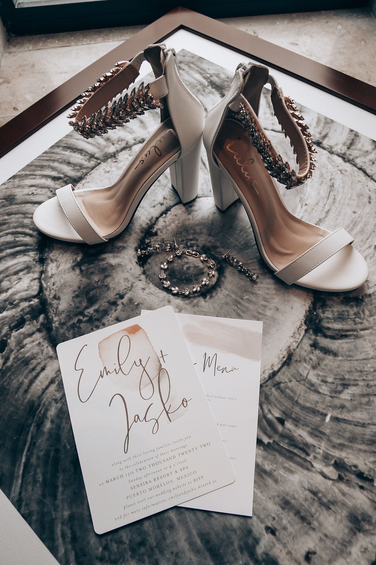 Wedding invitation cards displayed alongside elegant white bridal shoes and a pearl necklace on a marble surface.